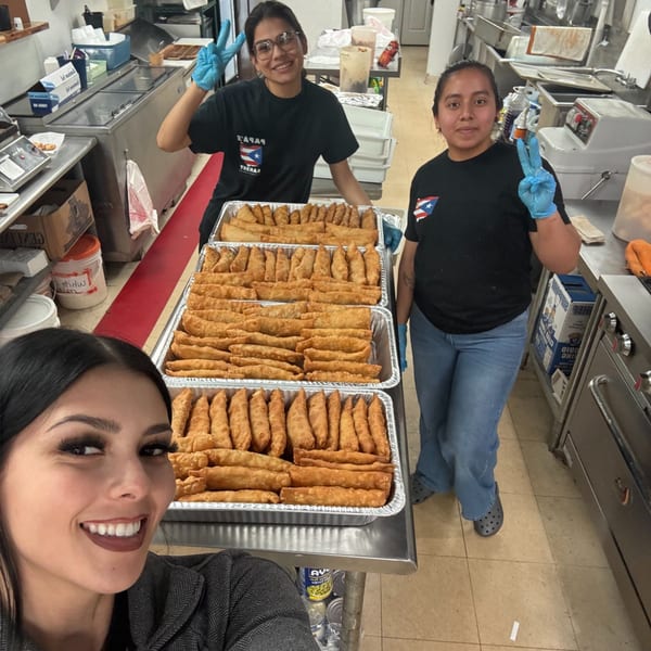 Three women in a kitchen with trays of Red Rican empanadillas, two making peace signs.